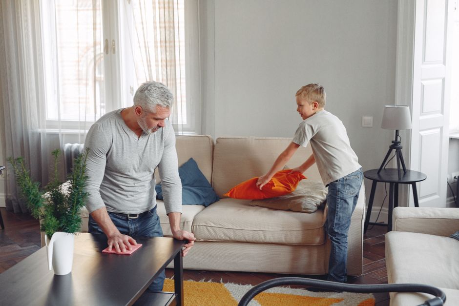 A father and son clean their living room, tidying up cushions and dusting tables.